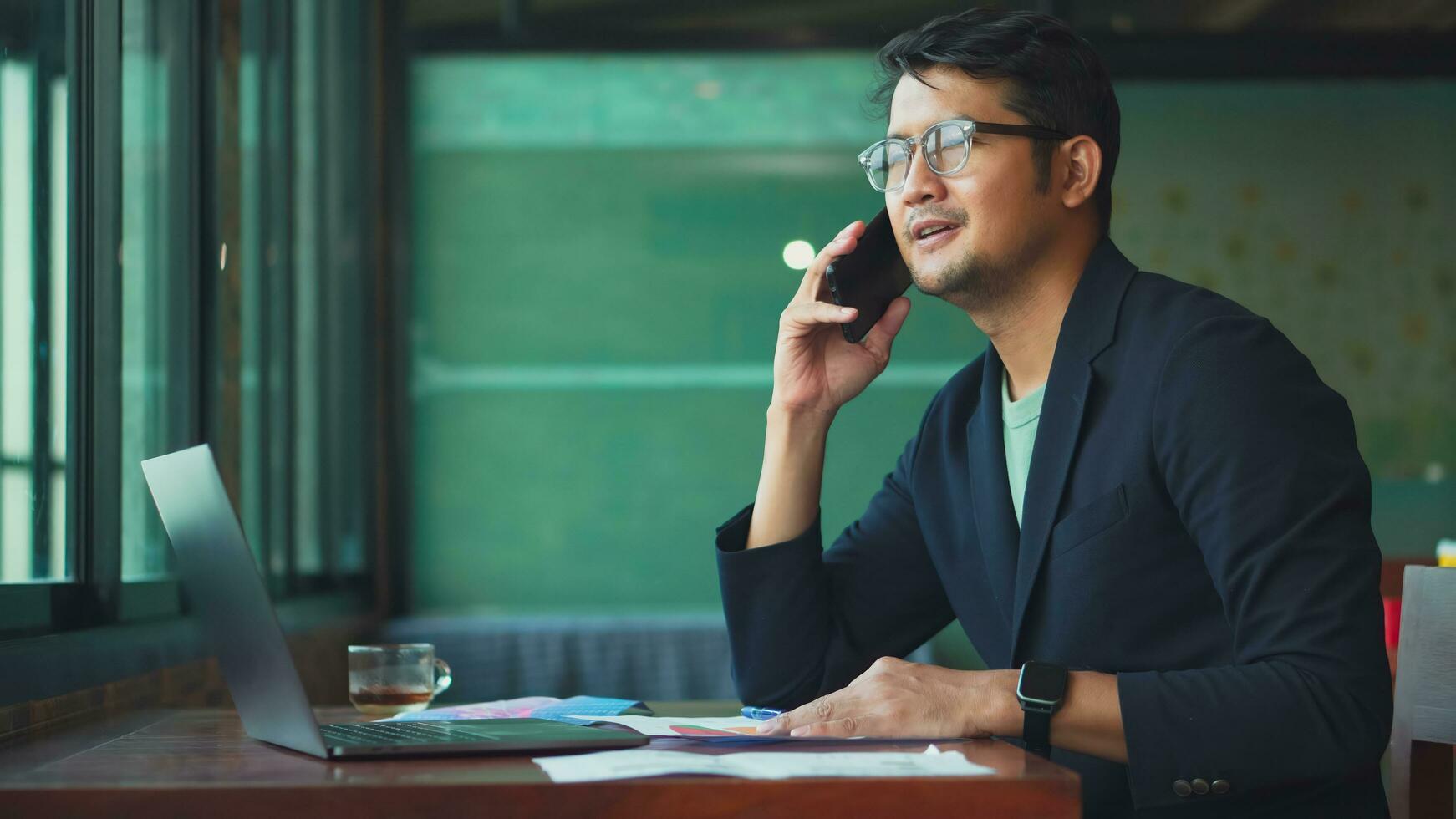 joven asiático hombre hablando teléfono y trabajo desde café con ordenador portátil computadora. foto