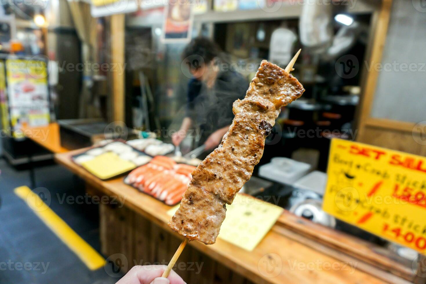 Closeup hand of tourist holding and eat barbecue Kobe beef wood stick on blurry barbecue Kobe beef shop at Kuromon market, Osaka, Japan. photo