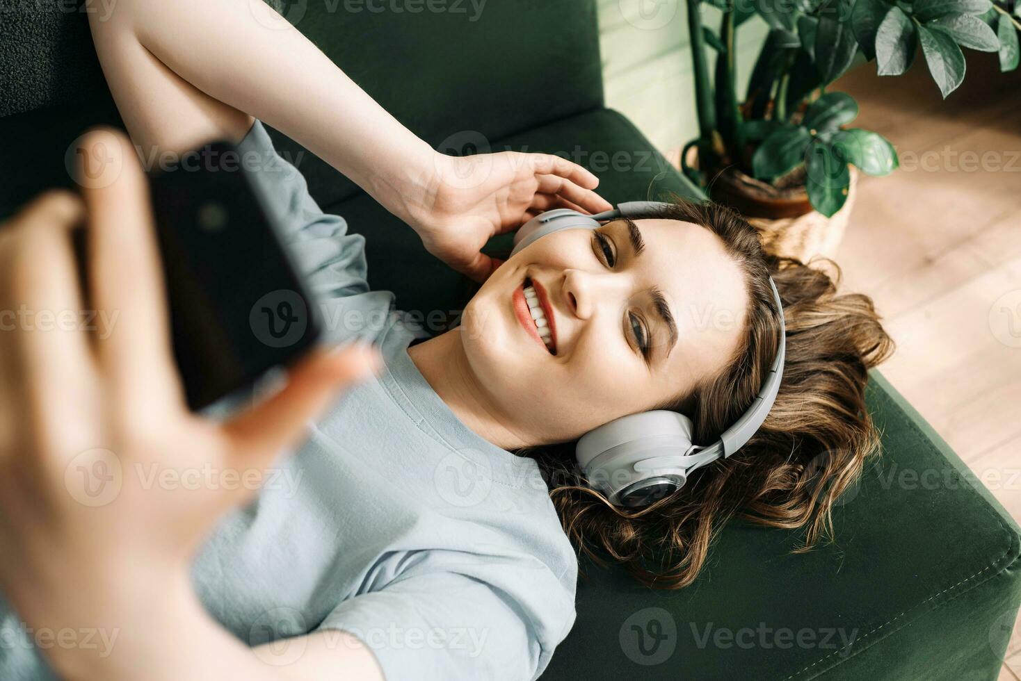 A smiling young middle-aged woman wearing wireless headphones listens to music while lying on the couch with her smartphone. Contentment in Tunes. Smiling Woman Enjoys Music with Wireless Headphones photo