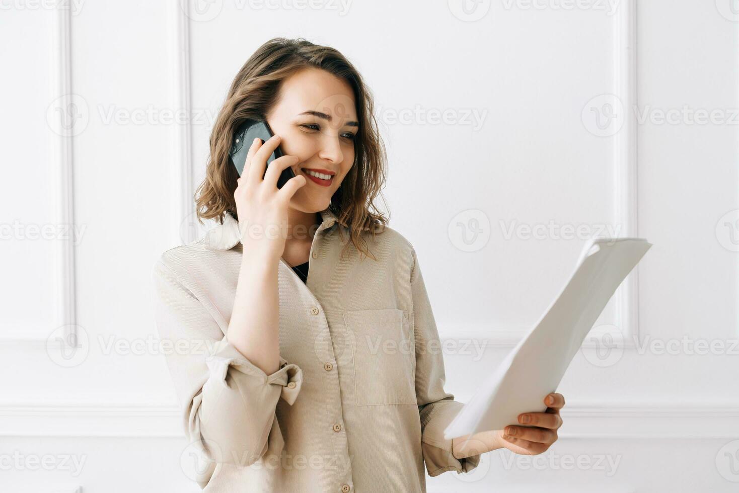 Smiling Millennial Woman Engaged in Phone Call, Discussing Documents with Client. Smiling millennial woman calling by smartphone, talking with client, looking at documents photo