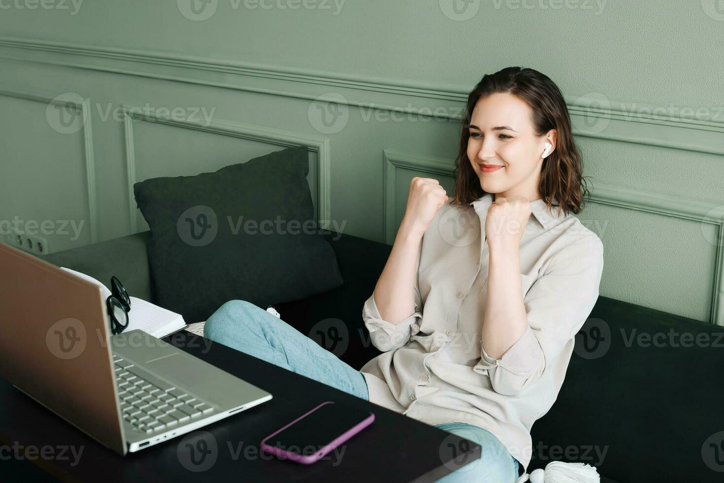 victorioso y celebrando. sonriente mujer gesticulando en un exitoso vídeo charla en su ordenador portátil. contento mujer en lentes es radiante como ella comunica en su computadora portátil, señalización victoria y éxito. foto