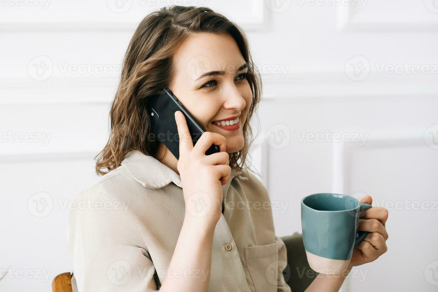 Happy Young Woman Enjoying a Phone Call While Savoring a Drink in Leisure Time. Joyful Young Woman Talking on Phone, Enjoying a Relaxing Moment with a Cup of Drink in Her Free Time photo