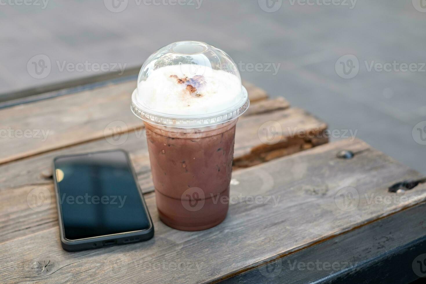 con hielo café, con hielo cacao en claro el plastico tazas cerca el negro teléfono inteligente en un antiguo de madera mesa en un relajante esquina café tienda. Copiar espacio foto