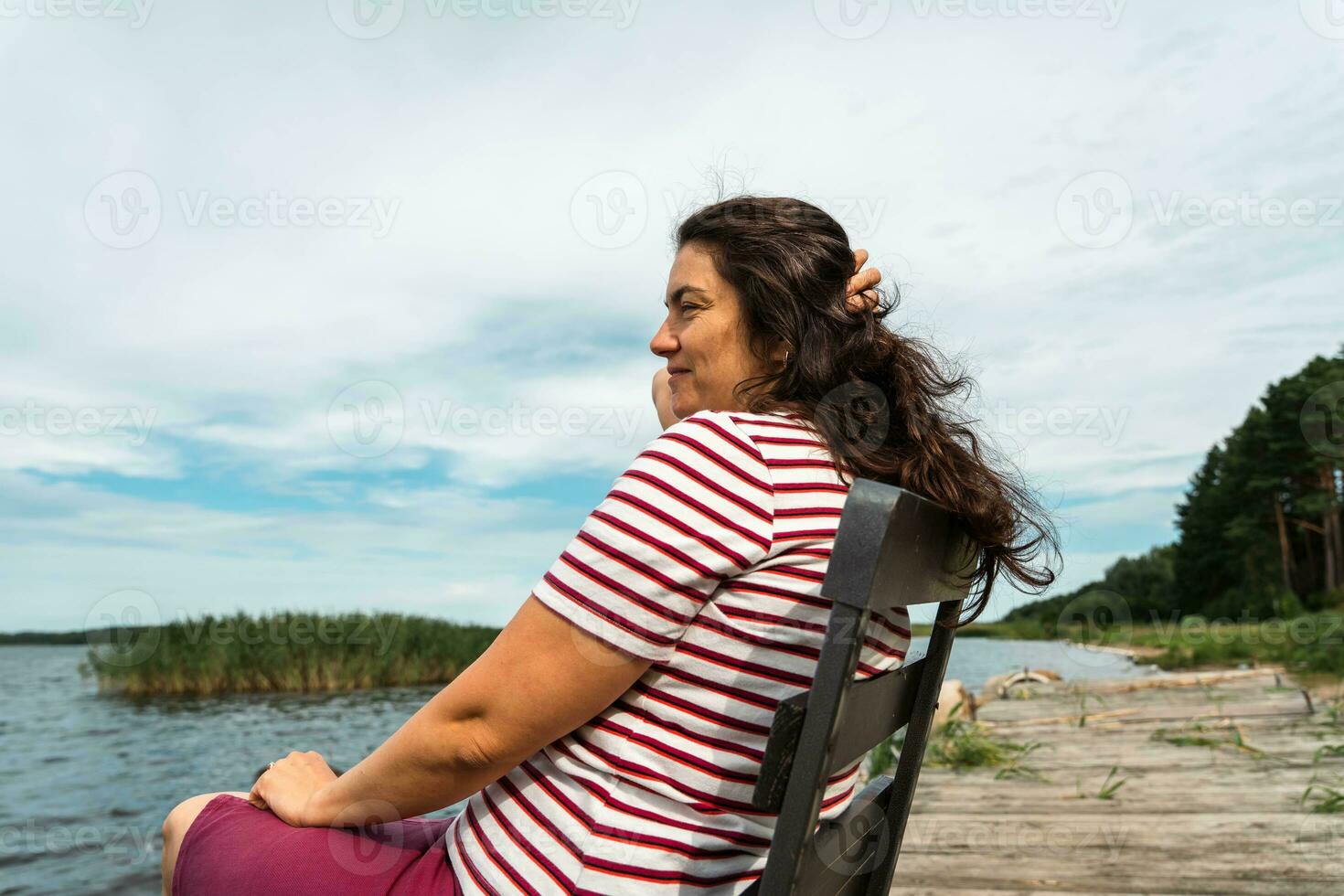 A long-haired woman is sitting alone on a chair on the shore of a summer lake, straightening her hair photo