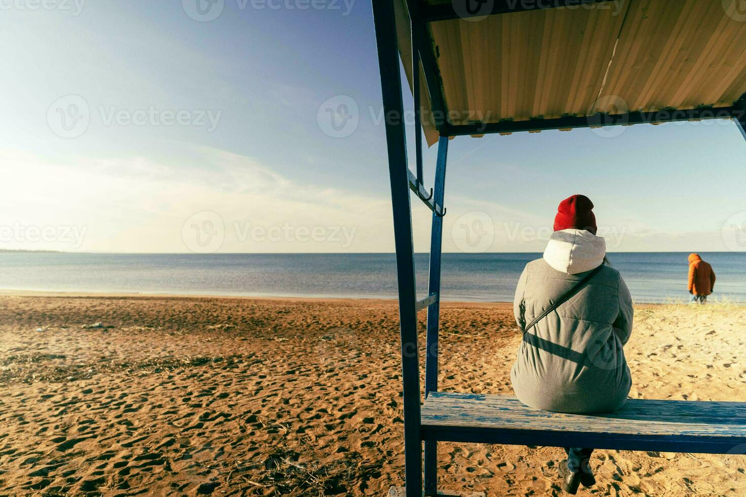 woman in an autumn jacket and hat is sitting alone on a bench on a deserted autumn beach on a sunny day photo