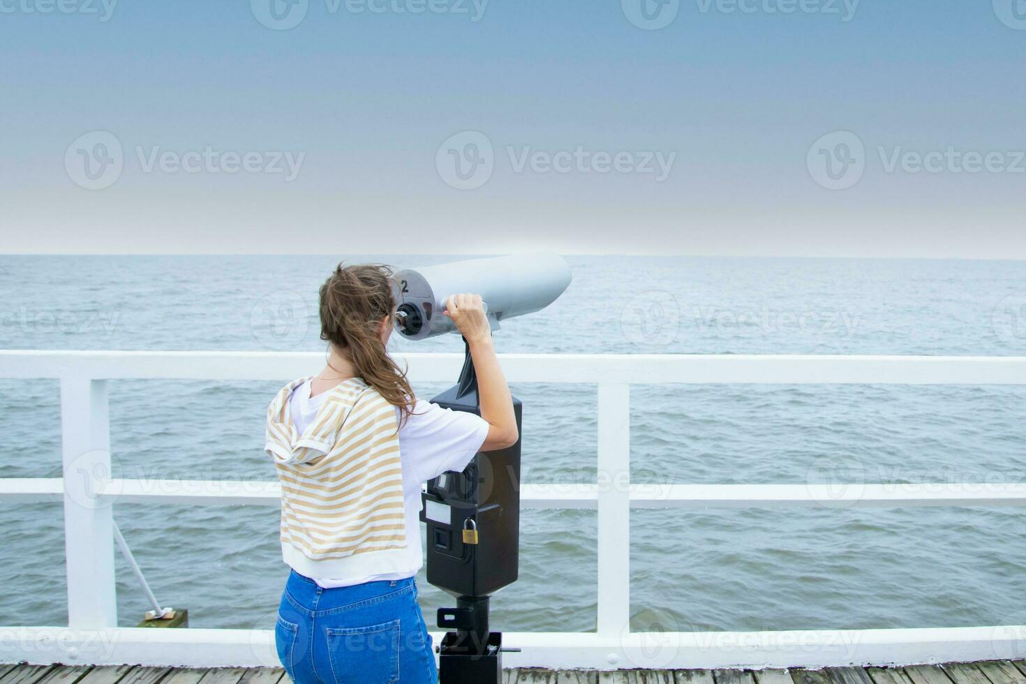 A young woman stands on the pier, looking through coin binoculars. rear view. baltic sea, cold weather photo