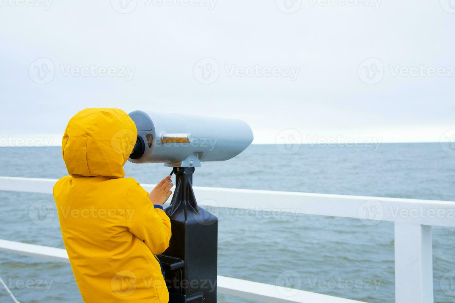 the boy is standing on the pier, looking through binoculars at the Baltic Sea. cold weather. grey sky photo