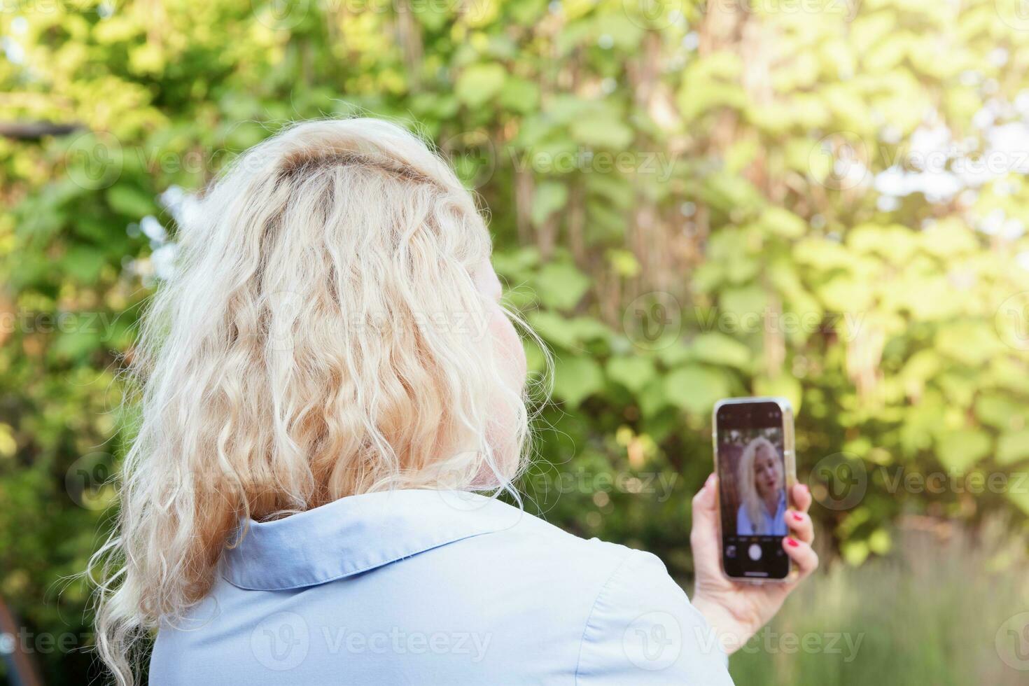 un linda joven mujer es sentado en un banco en el parque, utilizando su teléfono, tomando autofotos verano día. ver desde el atrás. foto