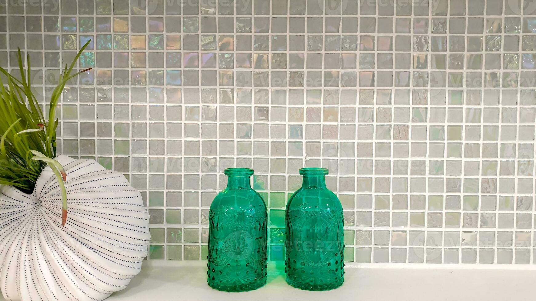 Ornamental plants in a giant shell shape 's ceramic vase with two glass bottles on  marble shelves and square tiles pattern wall background with space for texts. photo