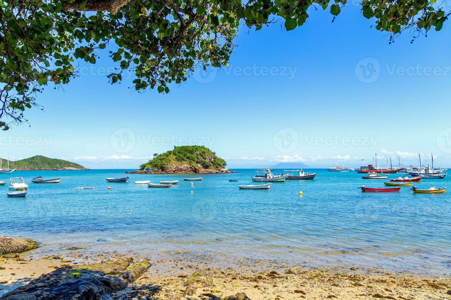 Seascape of Armacao Beach in Buzios, Rio de Janeiro, Brazil. Panoramic view of the bay. Boats and sailboats anchored in the sea. Tree leafs, clear water, blue sky, some rocks and a island. photo