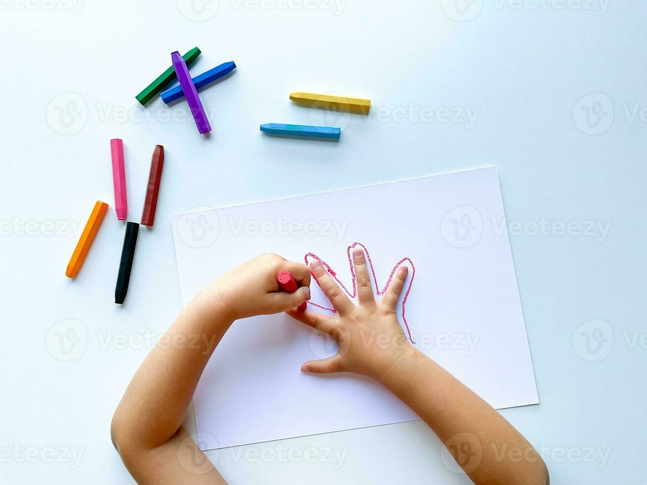 Childrens hands draw their hand with wax crayons on white paper, top view. photo