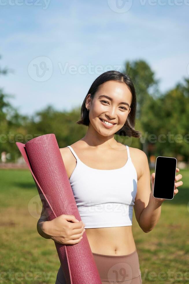 Excited fitness girl recommends application for sport and workout, shows phone screen, standing with rubber yoga mat in park after training session photo