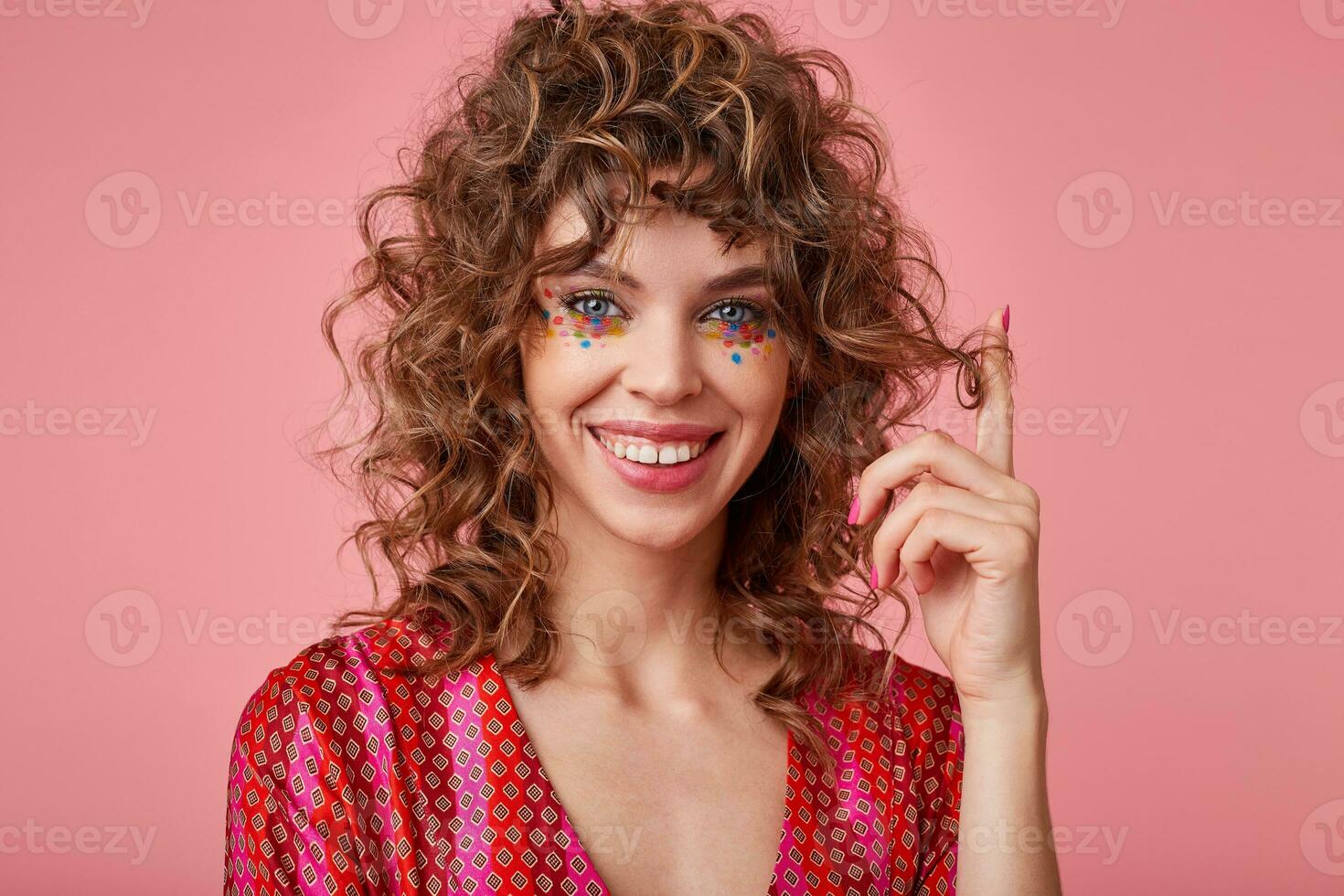 Young, positive woman with curls, plays with her hair, has multi-coloured dots on her face, stands over the pink background, smile at camera and looks joyful photo