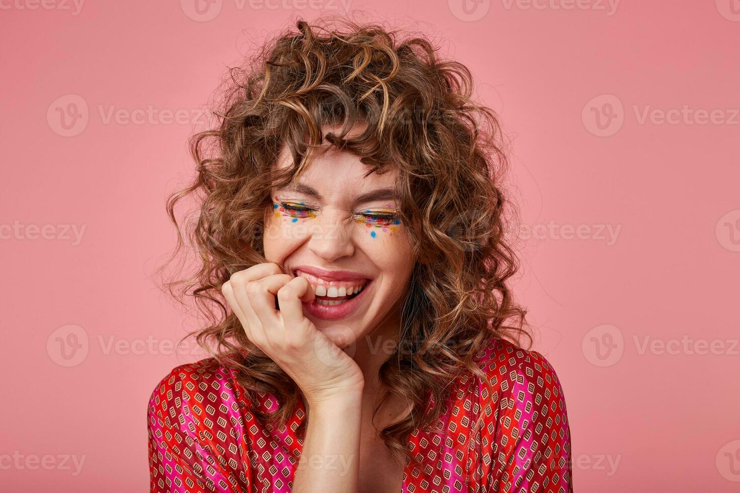 Close-up shot of joyful woman with curls, laughing at something with closed eyes, touching face with a hand, standing over the pink background. Positive emotion concept photo