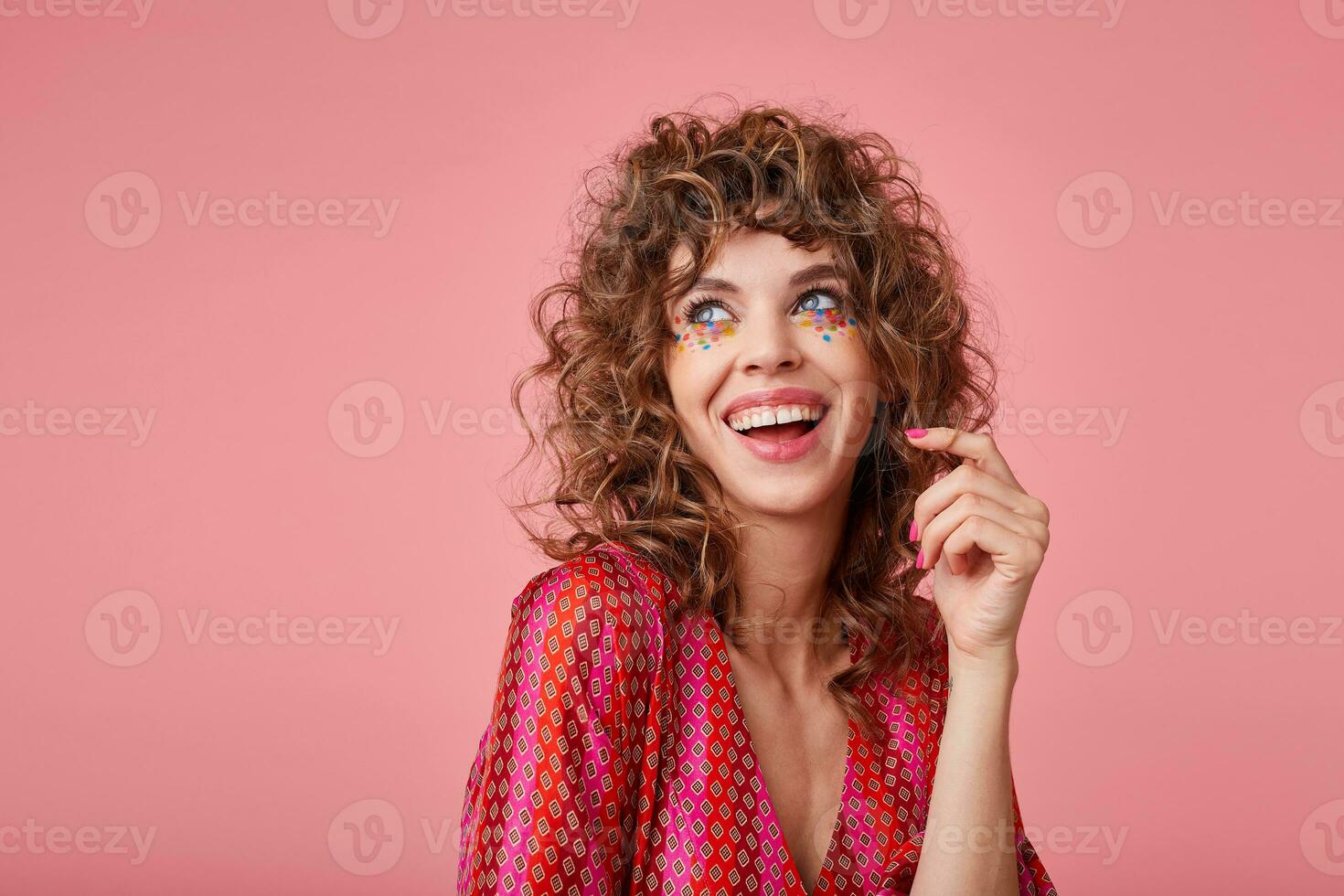 Beautiful young woman with curls laughing, being in a nice mood, standing over the pink background, wearing striped clothes with pattern, twisting a hair on a finger, looking away photo