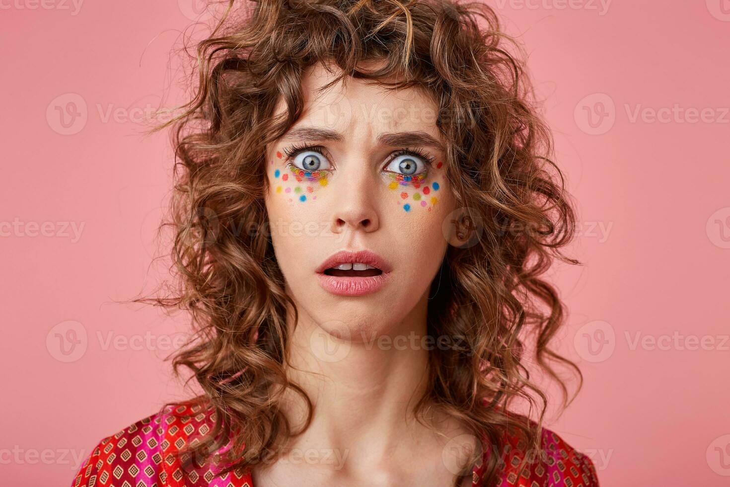Close-up shot of amased young woman with wide open eyes, looking at camera, isolated over pink background, wearing striped pink and orange clothes with pattern photo