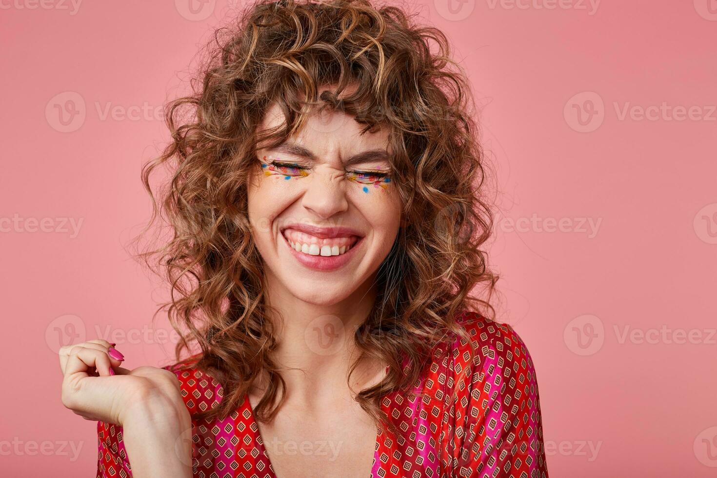 Pretty young curly woman smiles broadly, laughing at something with closed eyes, wearing striped clothes with pattern, standing over the pink background photo