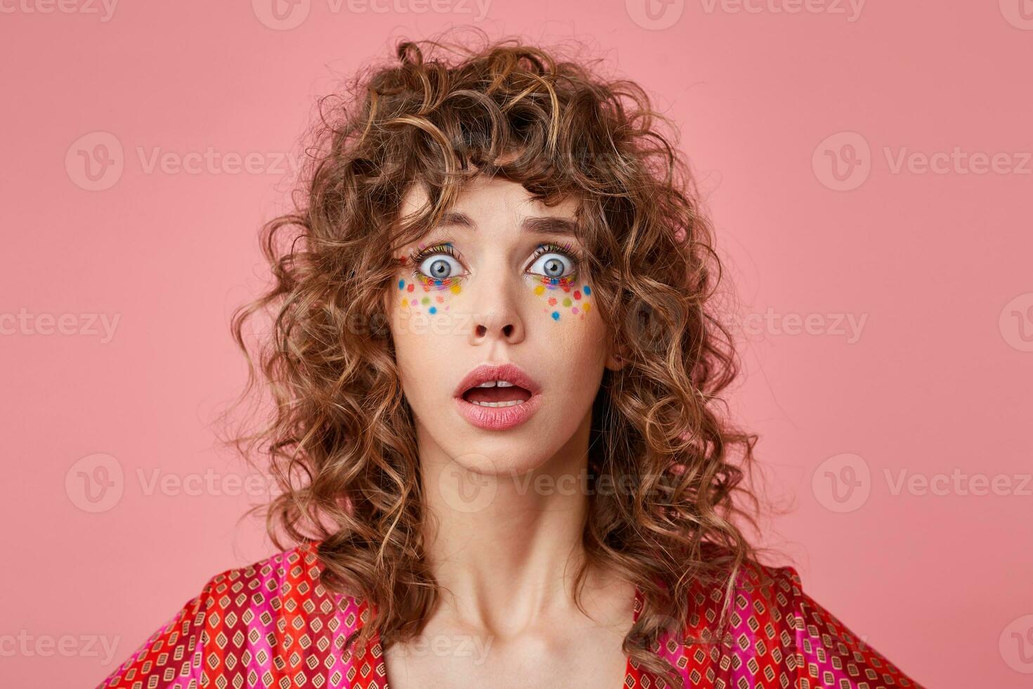 Portrait of shocked young curly woman with wide open eyes and mouth, isolated over pink background, wearing striped pink and orange clothes with pattern photo