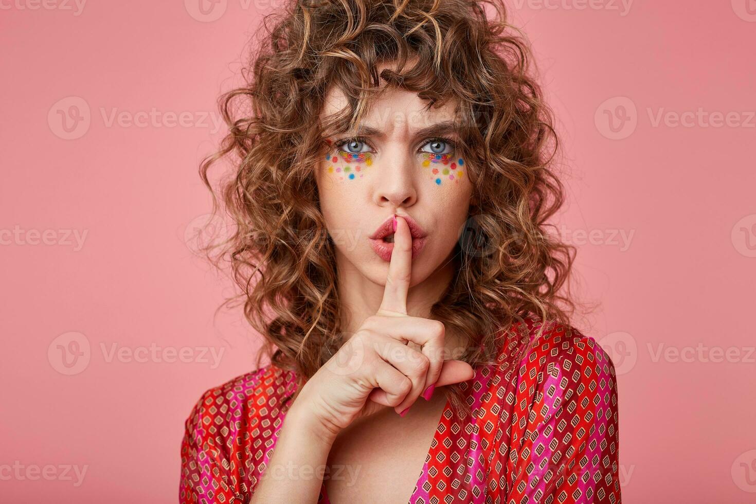 Close-up photo of serious curly young female with finger on her lips, urges to keep silence, to behave quietly, isolated over pink background