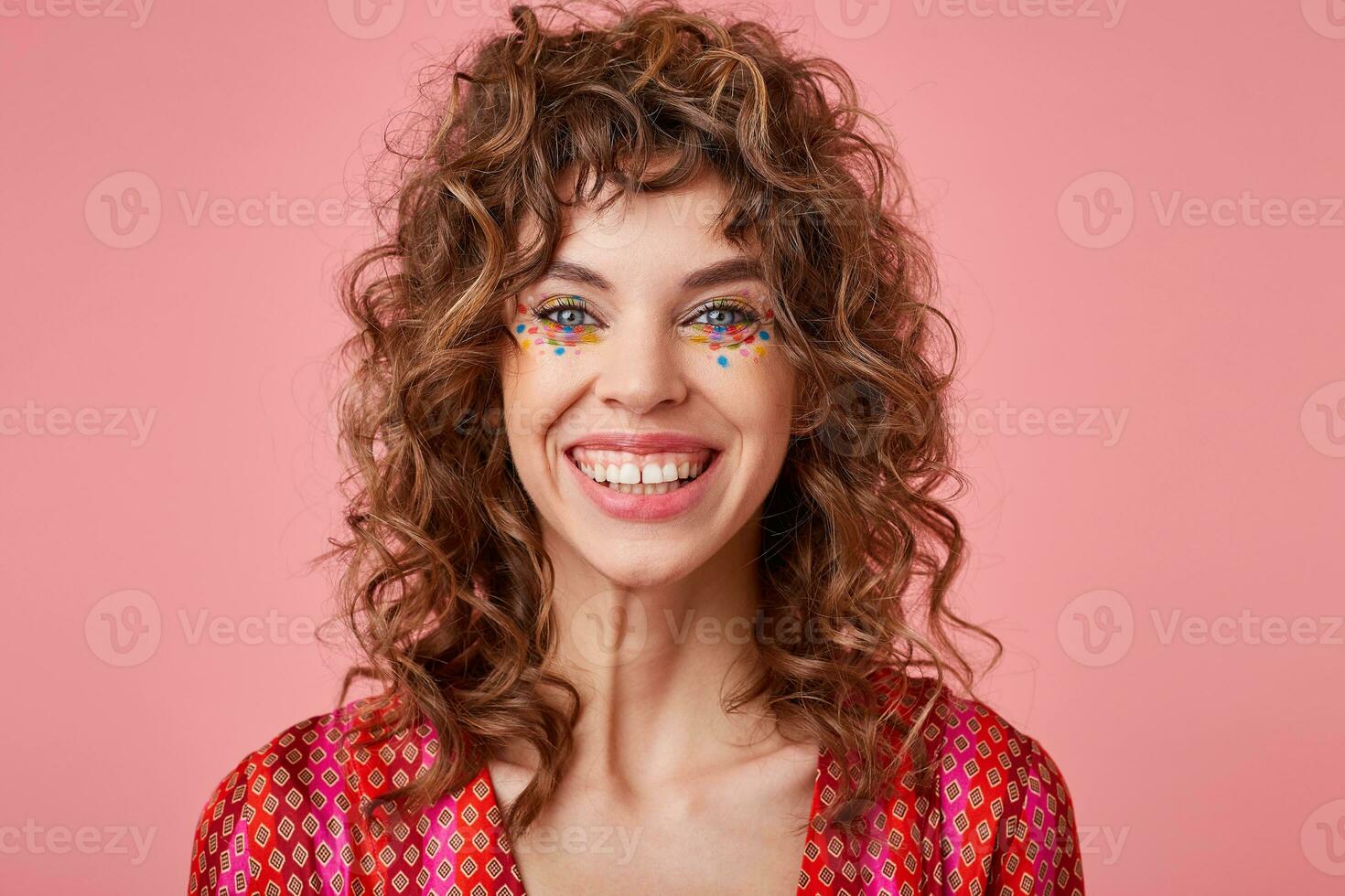 Portrait of beautiful young woman with curls, smiling cheerfully, showing her white teeth to camera, looking happy, standing over pink background photo