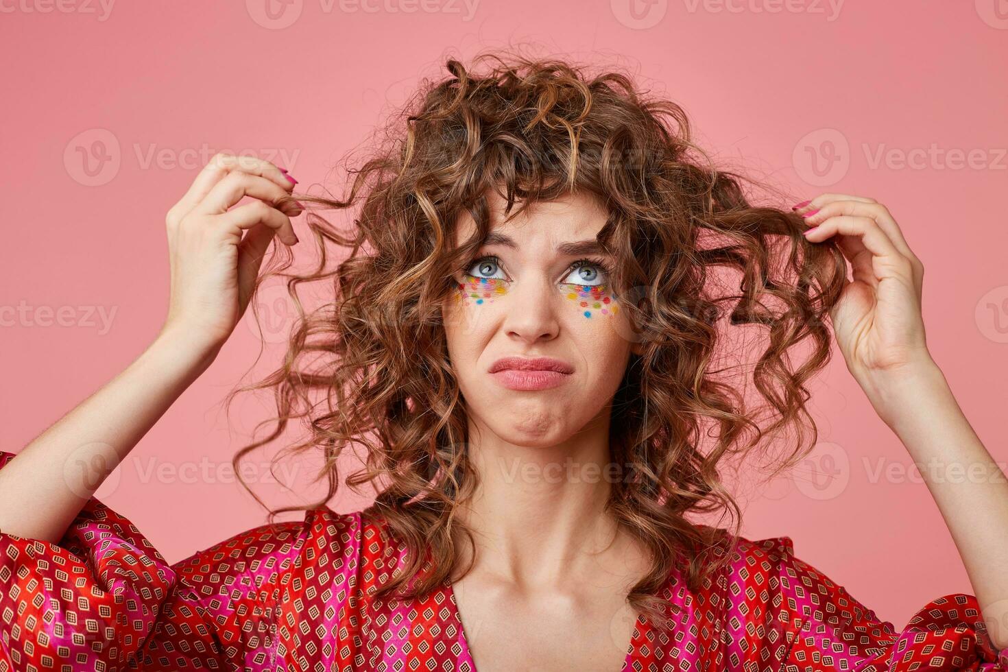 Close up shot of young woman looking up on pink background, pulling curly hair and being sad, making grimace, showing disappointment photo