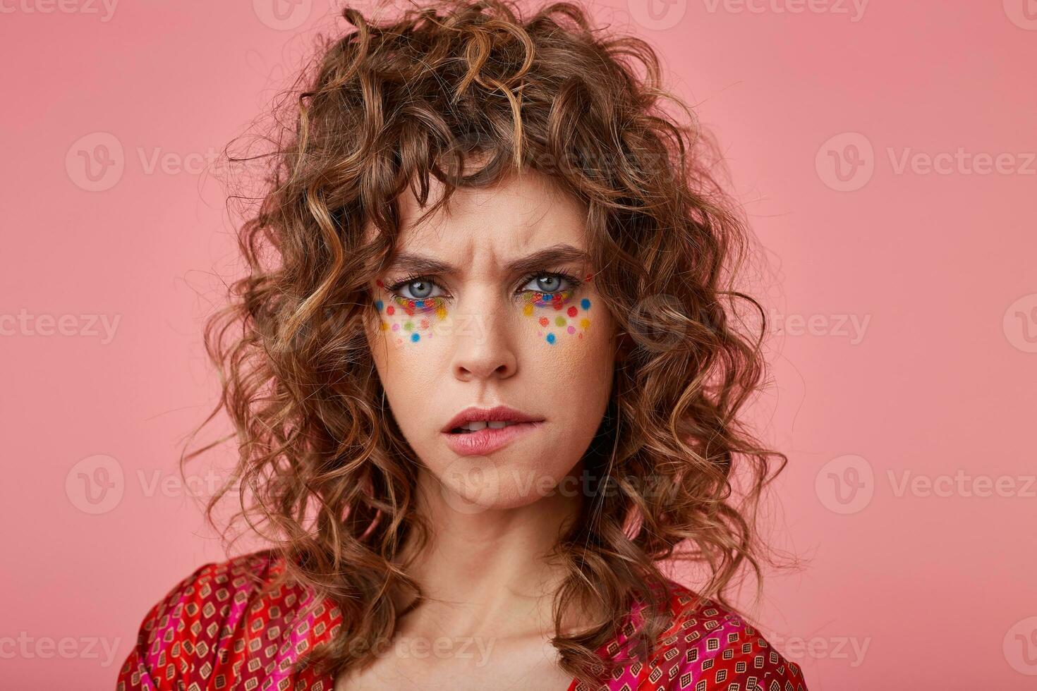 Young attractive woman frowning and biting her lip, looking serious and doubtful to the camera, standing over pink background, wearing striped pink and orange clothes with pattern photo