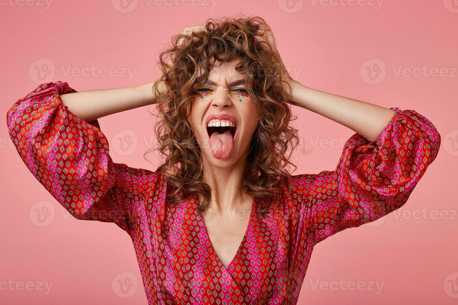 Portrait of excited young woman, screaming loud and sticking out tongue, covering ears with hands, standing over pink background. wearing striped pink and orange clothes with pattern photo