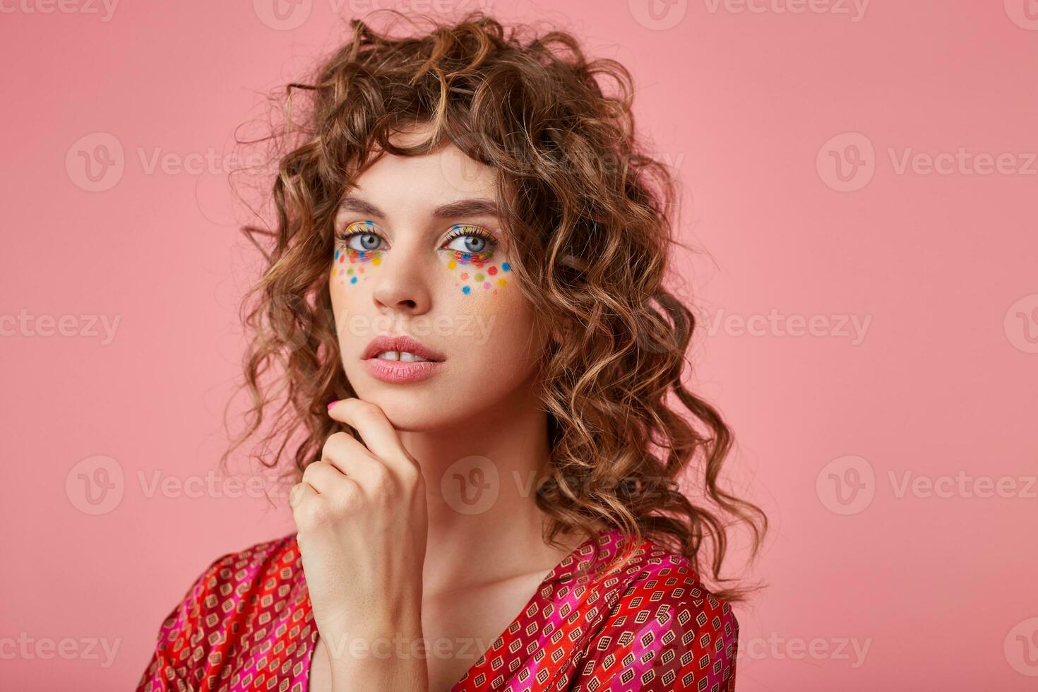 Young pretty woman over the pink background keeping hand under chin, wearing striped pink and orange clothes with pattern, looking calm to the camera with light smile photo