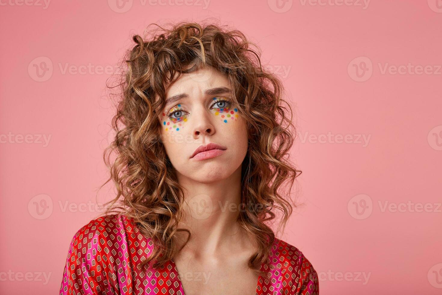 Indoor shot of unhappy young woman looking to the camera with sad face, showing gloomy expression, standing over pink background photo