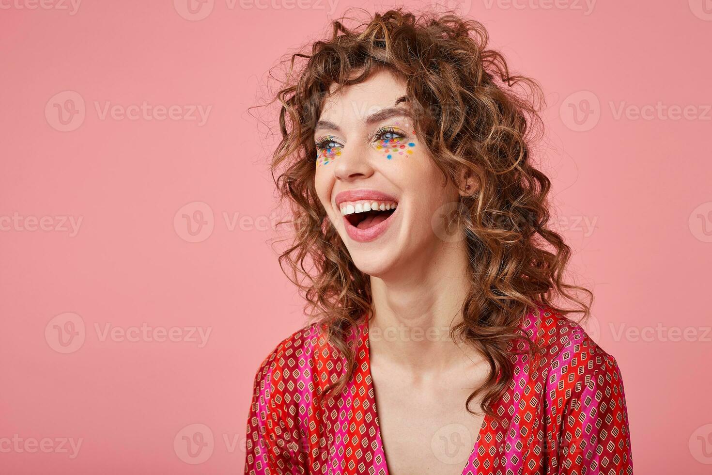 Pretty young curly lady looking away, being in nice mood, laughing, standing over pink background, wearing striped pink and orange clothes with pattern. Positive emotions concept photo