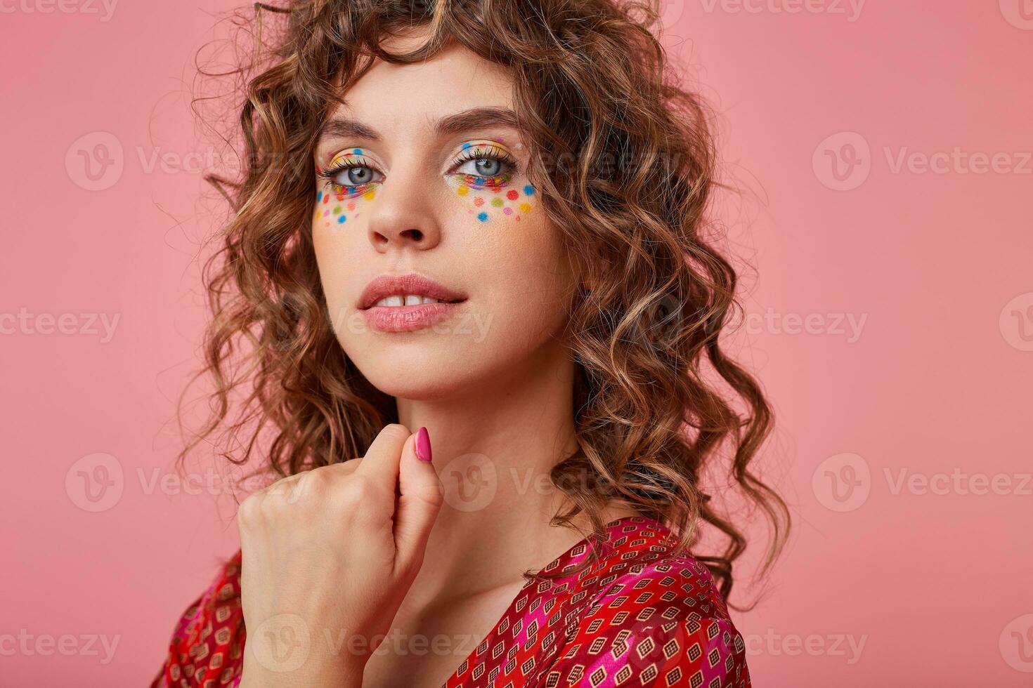 Sensual portrait of young female with curls, smiling softely and keeping hand under the face, isolated over pink background, wearing striped pink and orange clothes with pattern photo