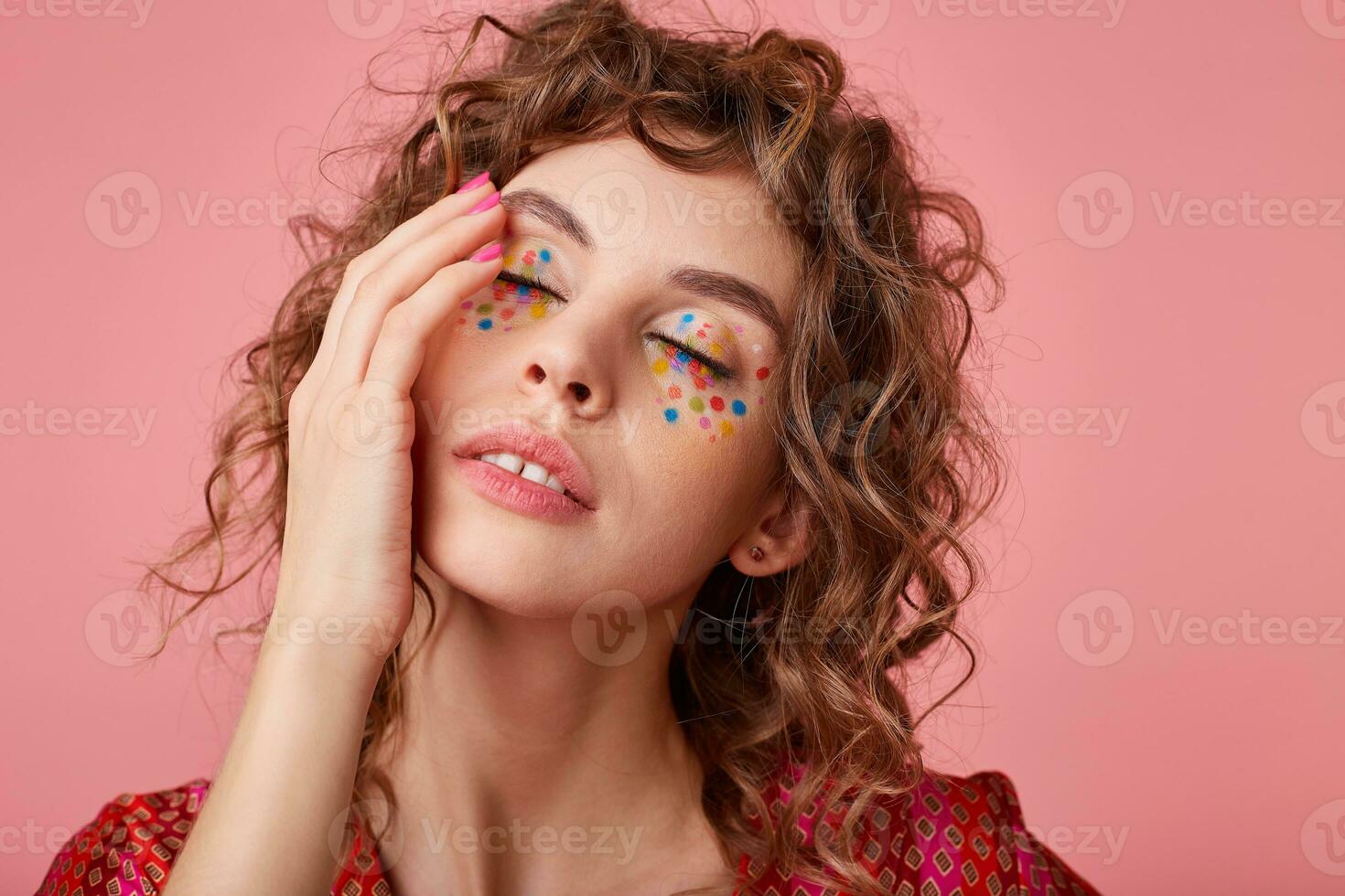 Dreamy cute young woman with romantic smile, touching gently her face, posing on pink background with eyes closed, wearing striped pink and orange clothes with pattern photo