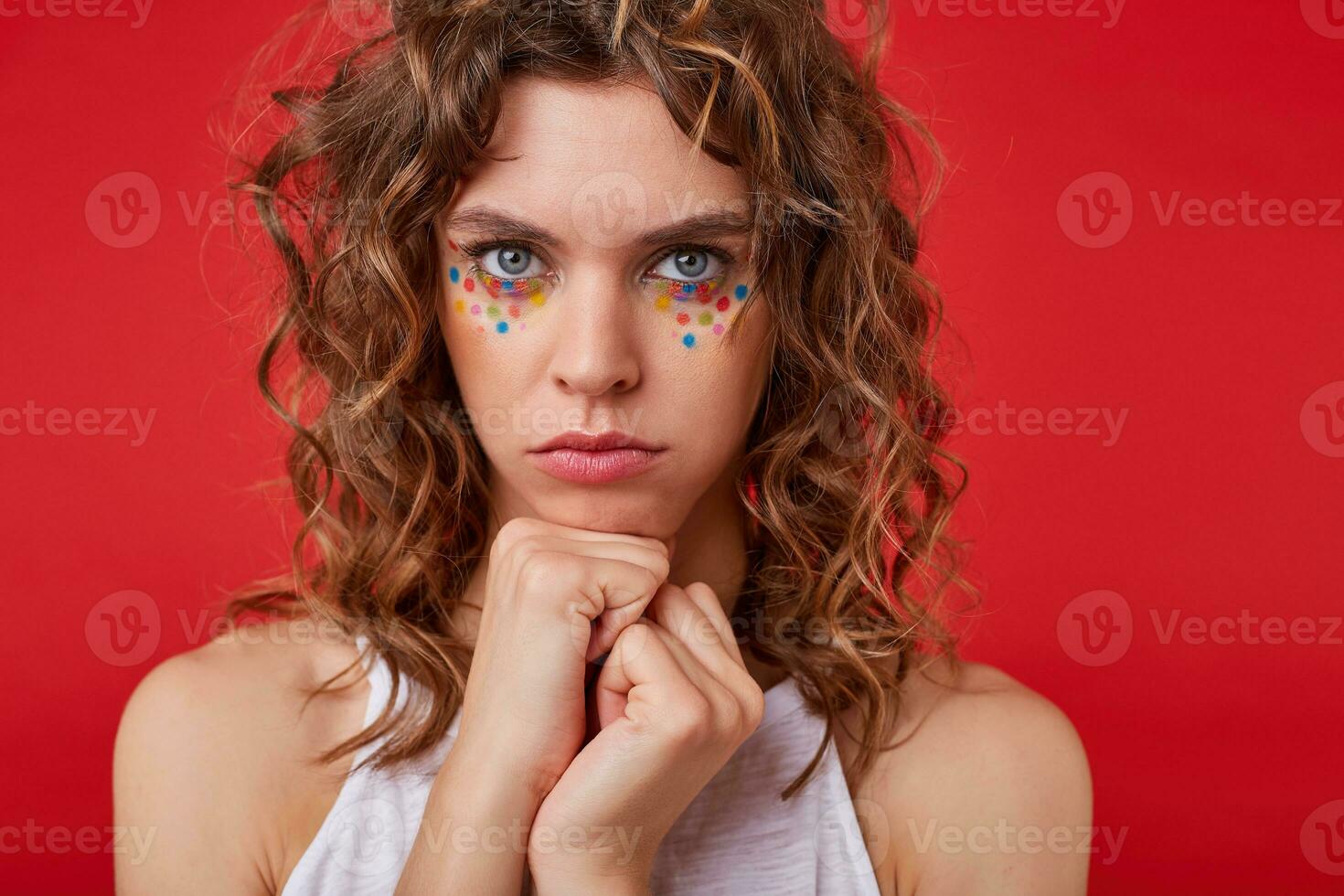 Pretty young woman with light-brown curly hair standing over the red background, looking offended and confused, clenching fists near to face photo