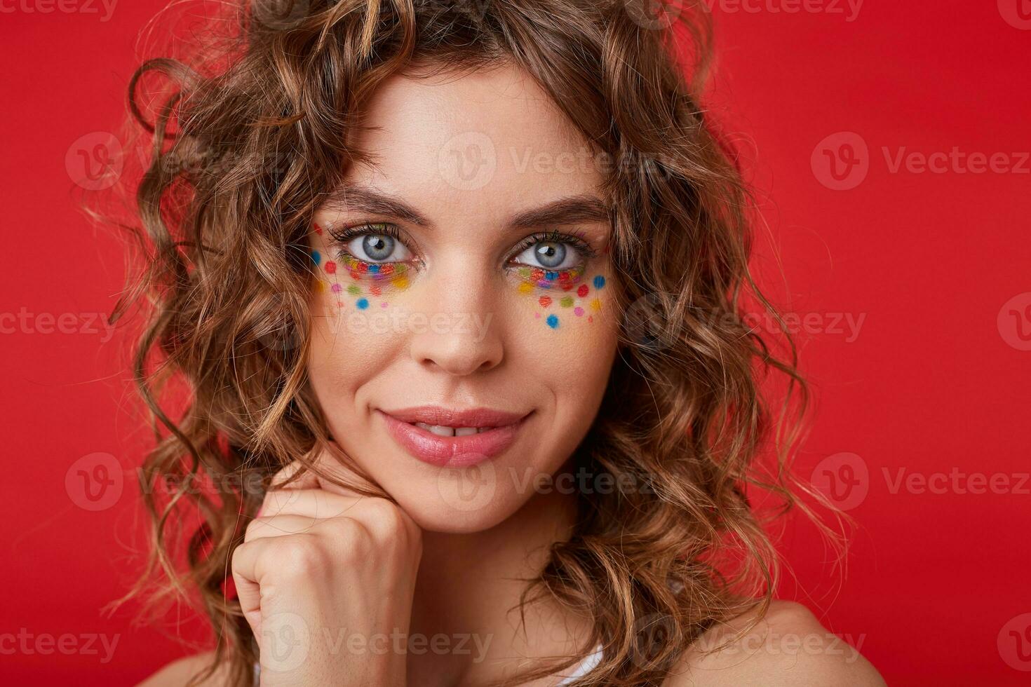 Close-up photo of cute young woman, smiling lightely, touching gently her cheek, looking to the camera, keeping hand in fist, posing on red background