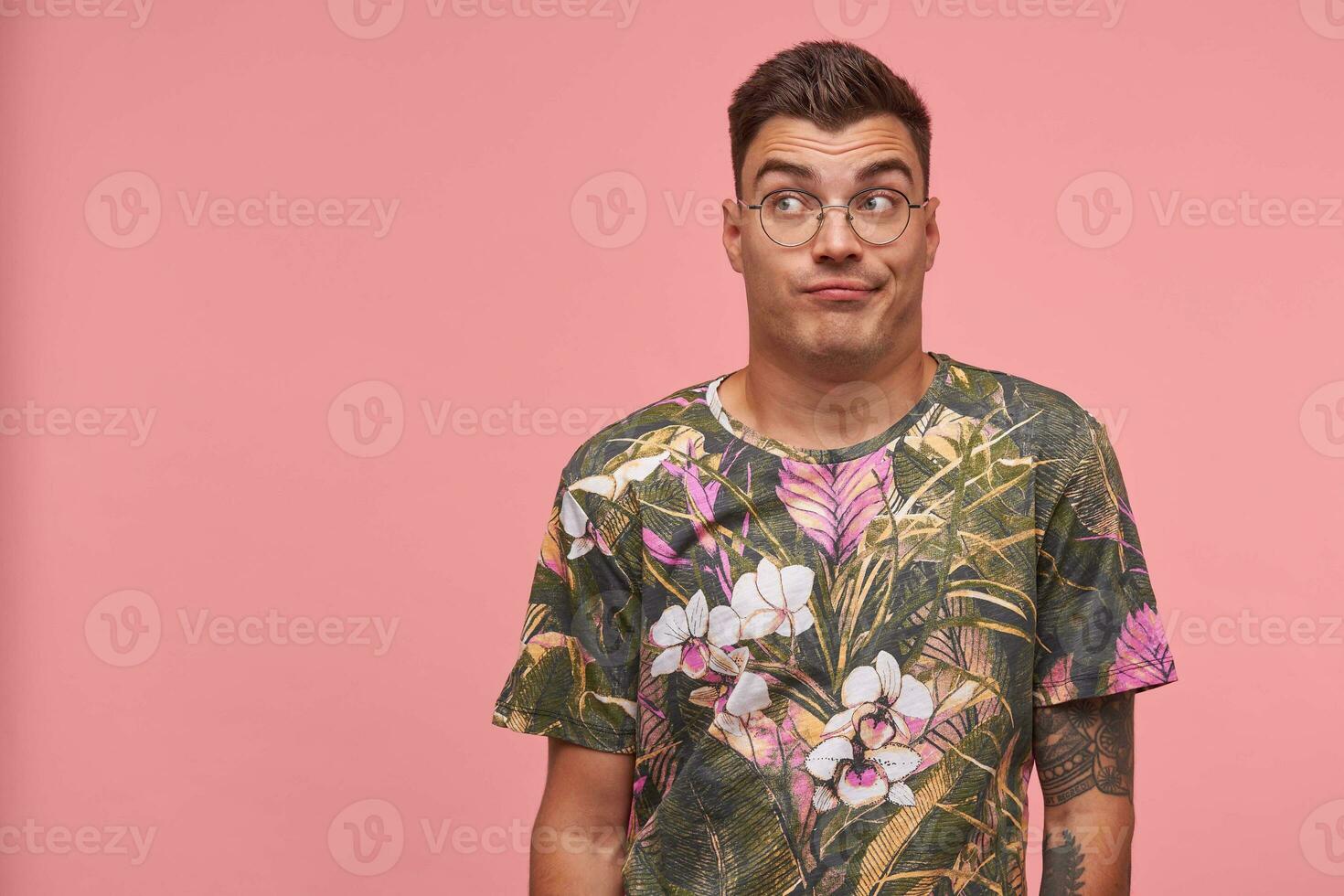 Young attractive man standing in front camera over the pink background wih raised eyebrows, looking away, wearing glasses and printed t-shirt, seems bored and indifferent photo