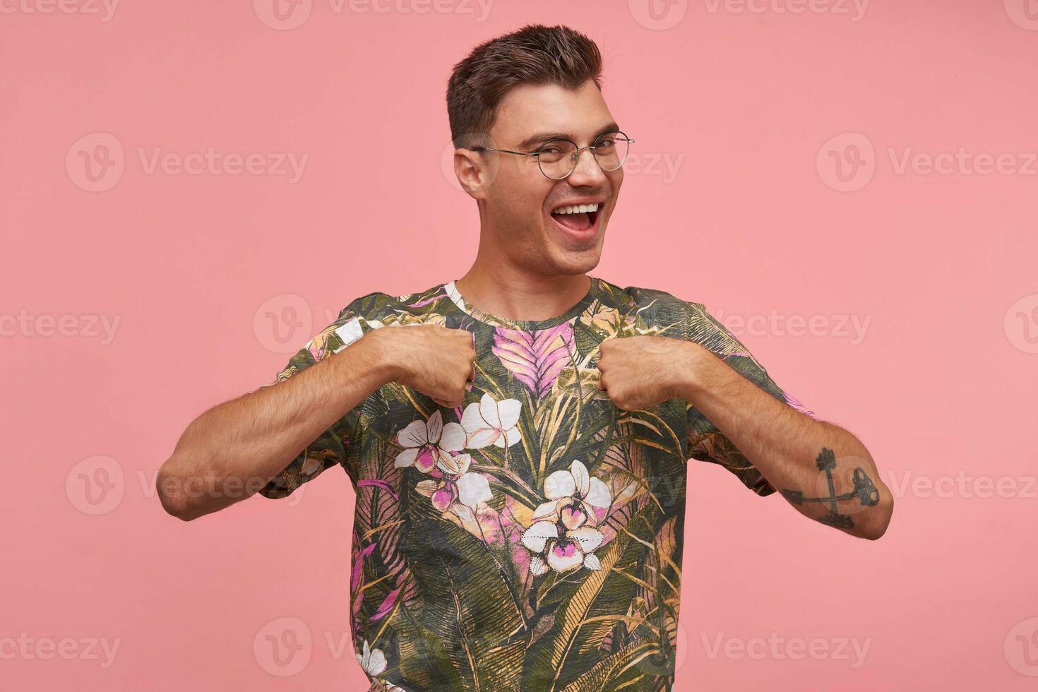 Pleased and smiling guy with hands on his chest looking at camera, wearing glasses and printed t-shirt, standing over pink background, positive emotions photo