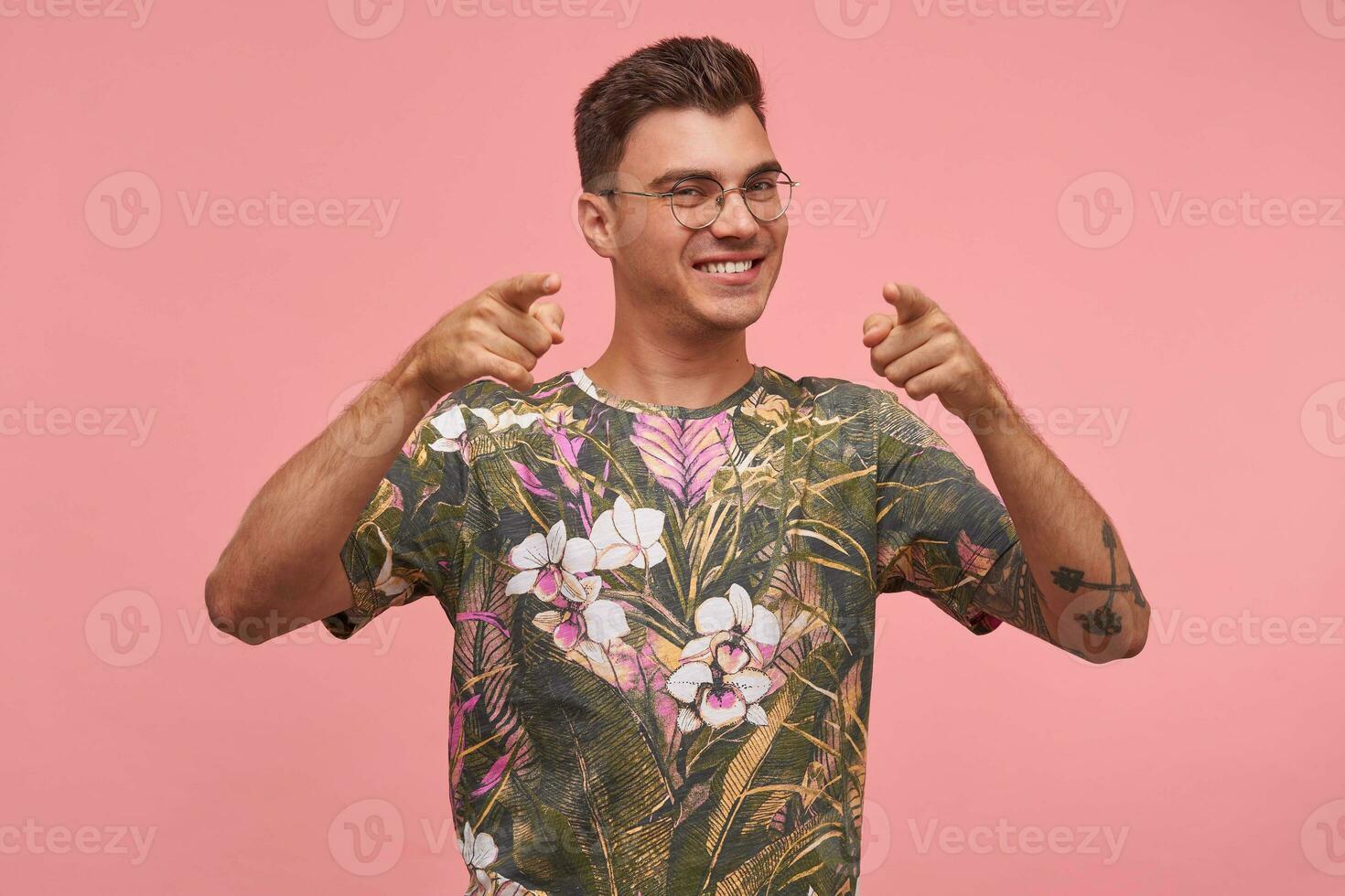 Good looking young man smiling broadly and pointing fingers at camera, wearing glasses and printed t-shirt, standing over pink background, being glad and joyful photo