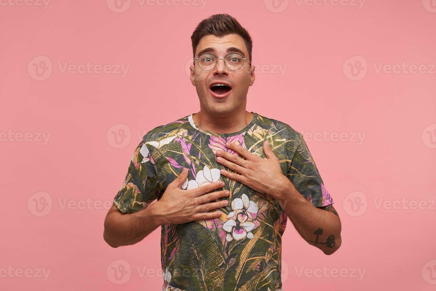 atractivo de pelo corto joven hombre con palmas en su cofre en pie terminado rosado fondo, vistiendo de flores camiseta y anteojos, mirando a cámara sorprendido foto