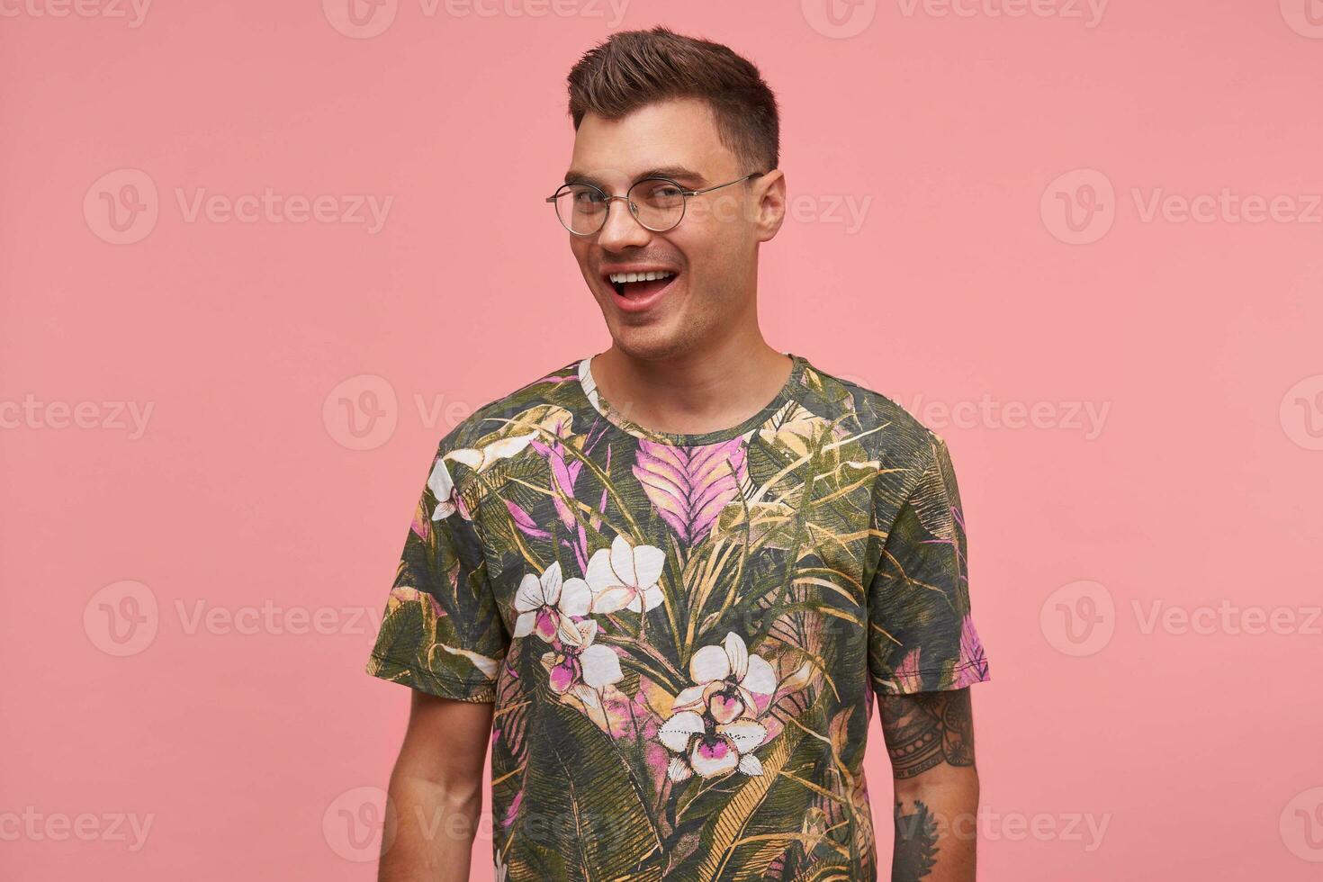 Young pretty guy looking at camera with wide smile, being positive and happy, wearing flowered t-shirt and glasses, standing over pink background photo