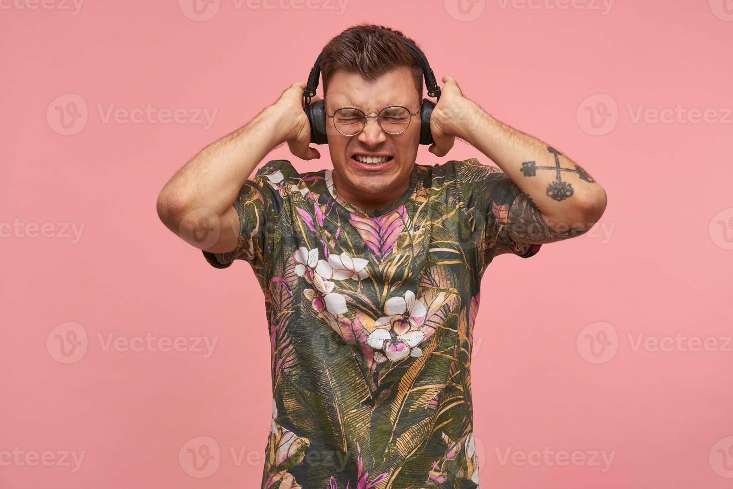 Young tattooed guy in flowered t-shirt listening music too loud, covering ears, squinting and biting the bullet, posing over pink background photo