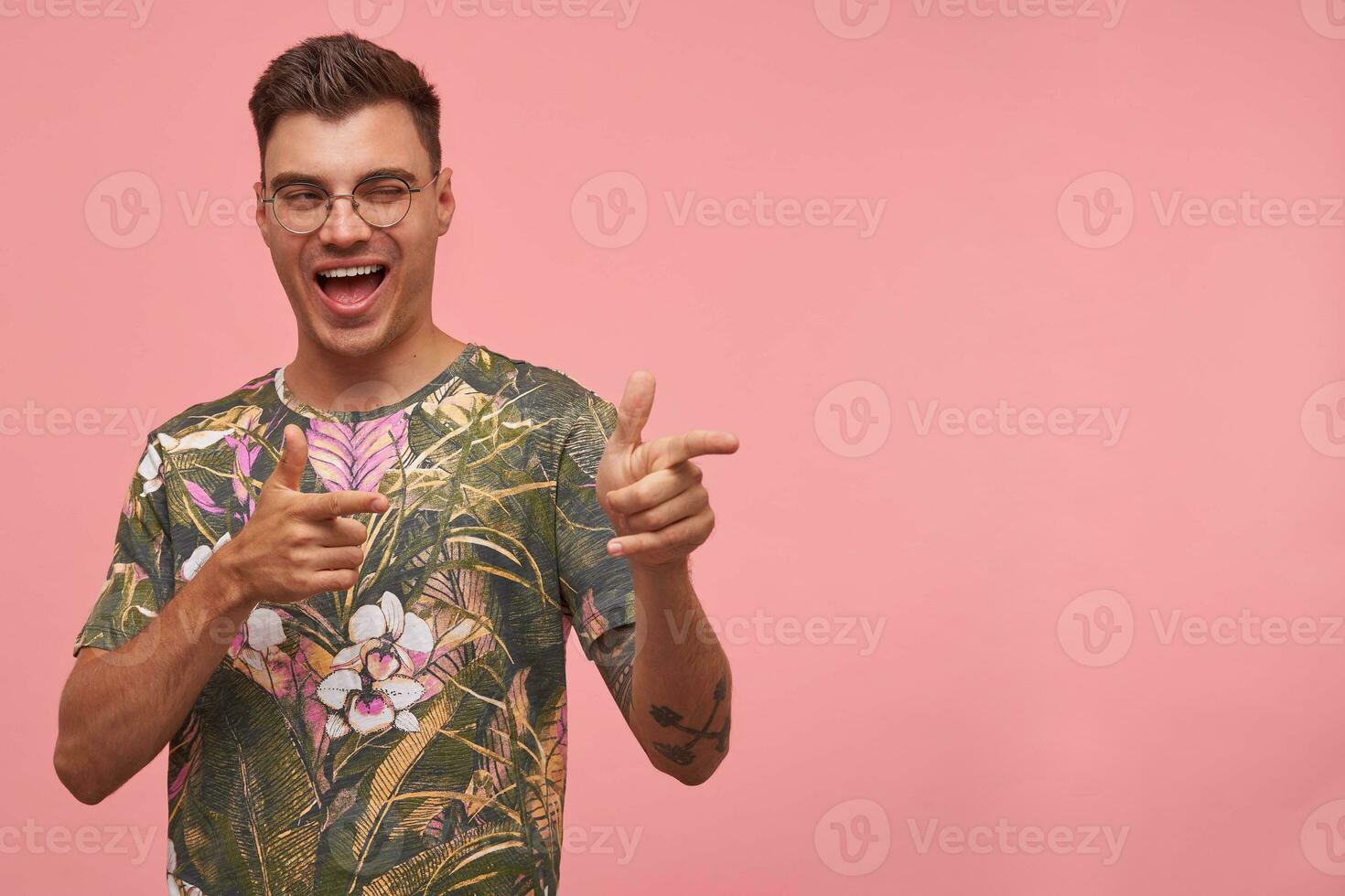 Indoor shot of enthusiastic attractive guy with glasses, indicating with fingers aside, giving wink, isolated on pink background photo