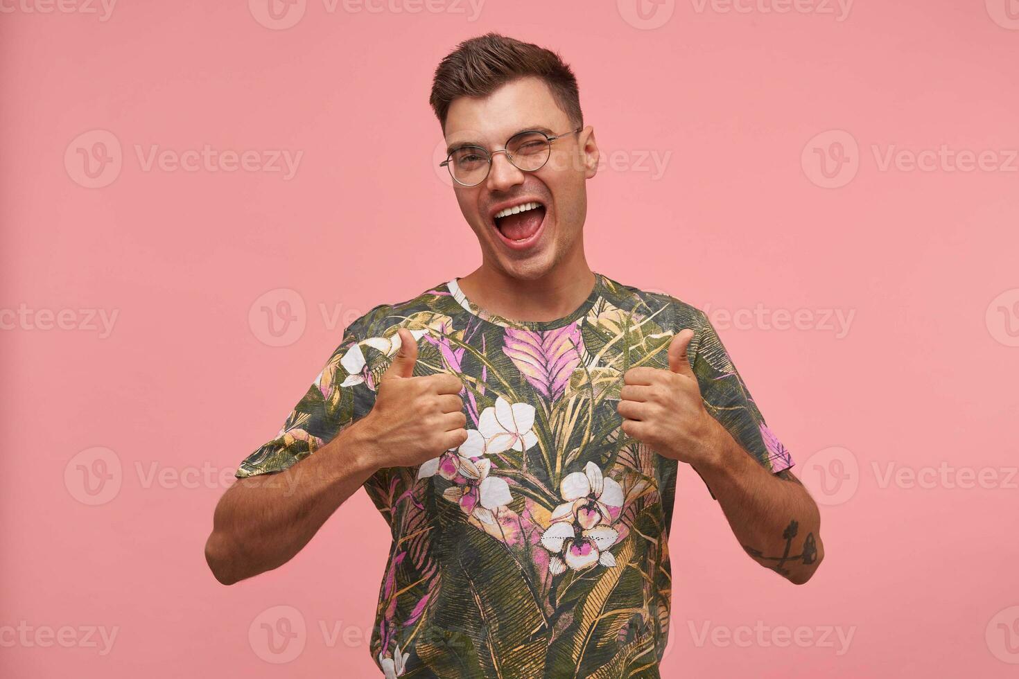 retrato de contento joven hombre vistiendo de flores camiseta, pulgar arriba y parpadeo a cámara, aislado terminado rosado fondo, disfrutando vida foto