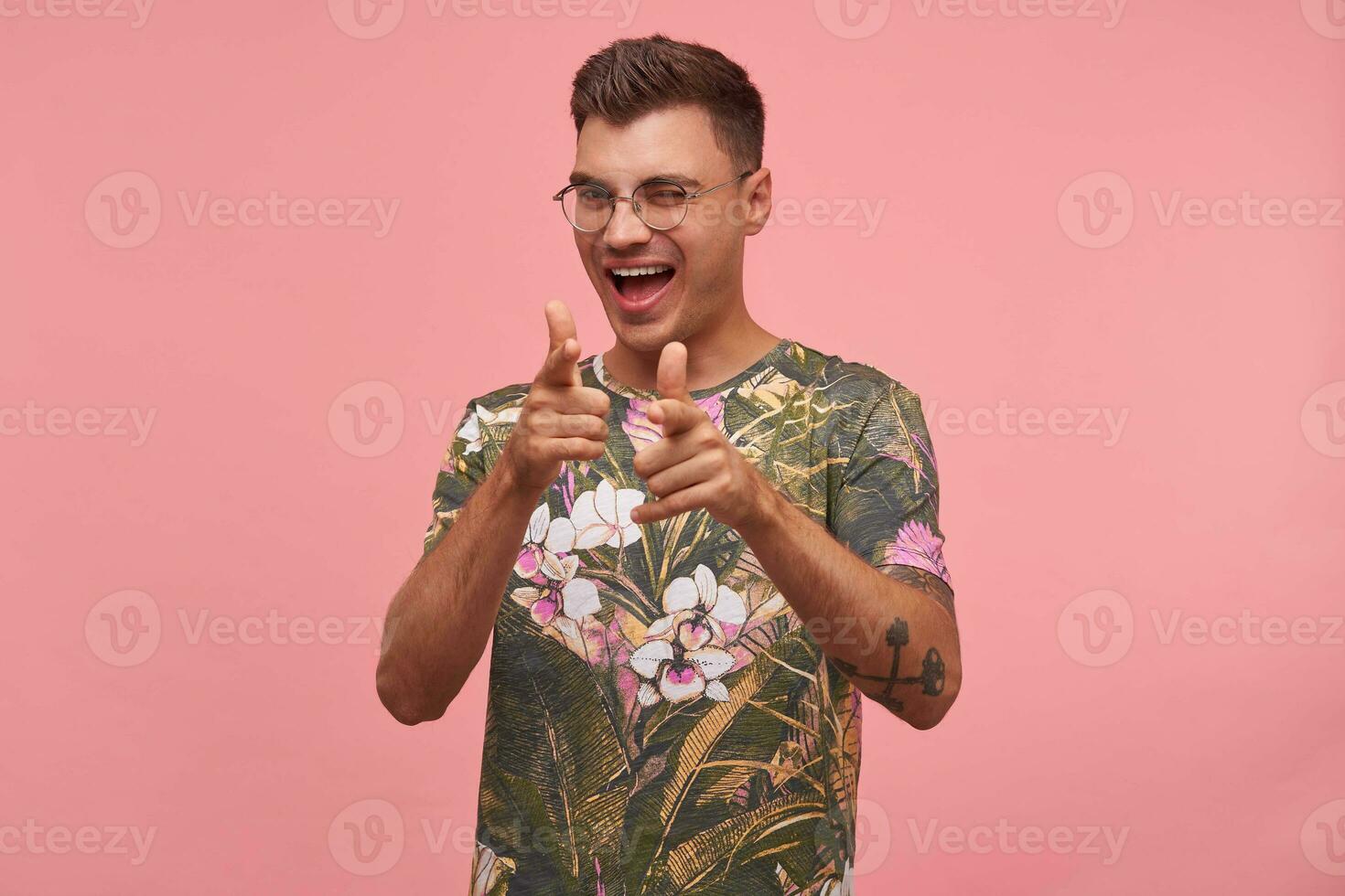 Portrait of a cheerful young man, pointing at camera, giving wink and smiling broadly, isolated on pink background, wearing flowered t-shirt and glasses photo