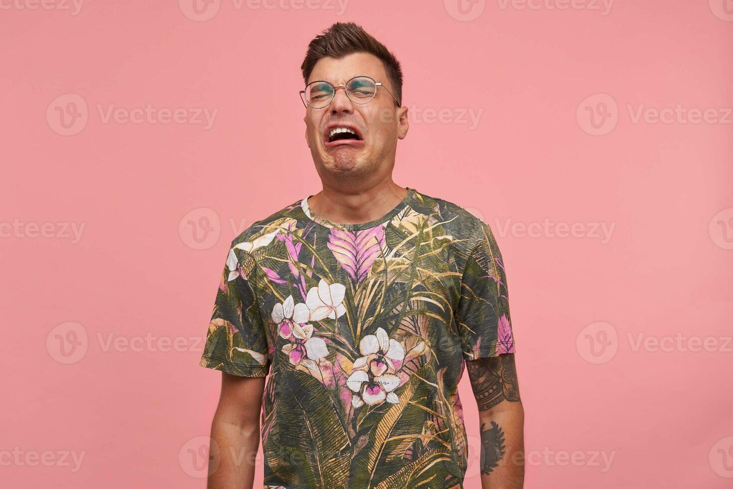 Close up of upset distressed young guy with closed eyes, crying a river over pink background, wearing glasses and flowered t-shirt. Sad emotions concept. photo