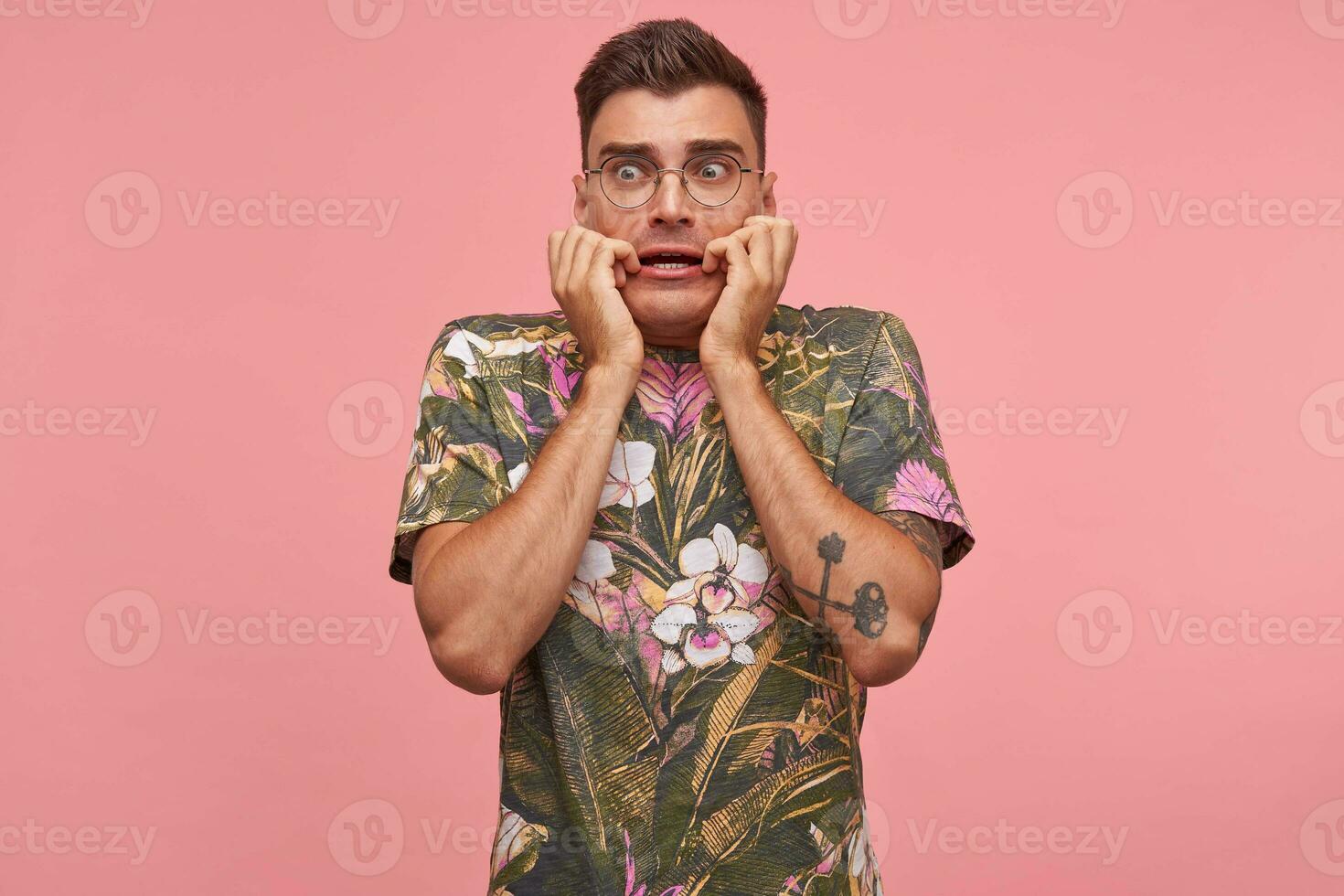 Young attractive man looking away with fear, keeping hands on face, standing with wide eyes opened over pink background, wearing glasses photo