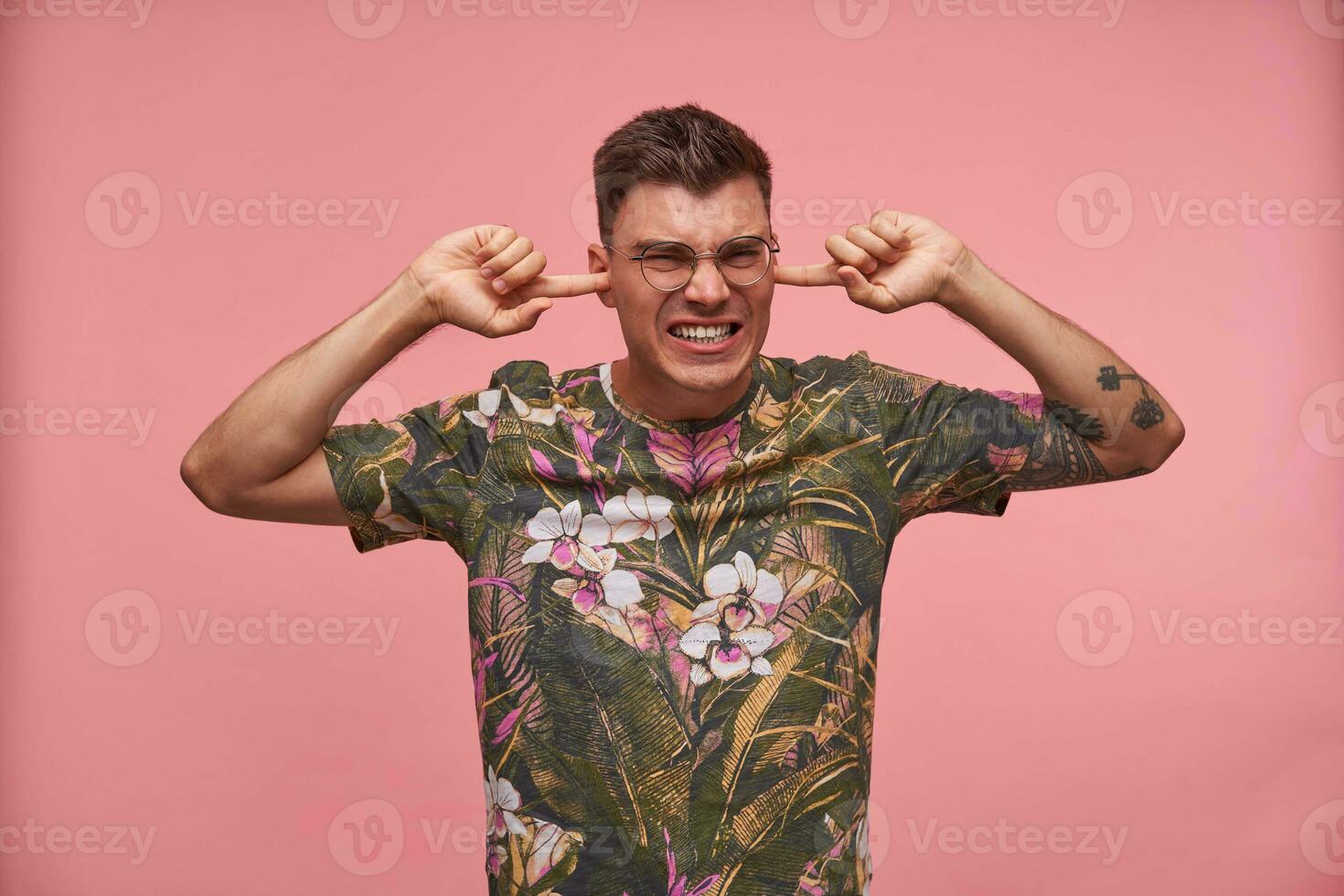 Stressed short haired young man with closed eyes pluging ears, avoiding loud sounds, ignoring something unpleasant, isolated over pink background photo