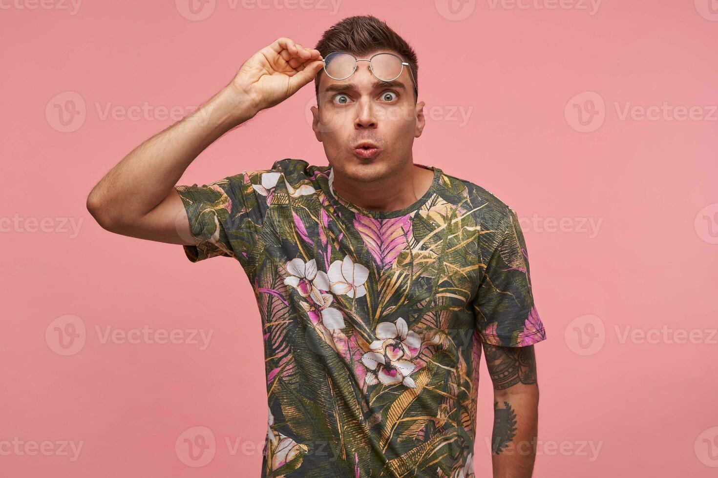 Suprised young short-haired man looking fearful to the camera with wide eyes open, pursing lips and holding his glasses, standing over pink background photo