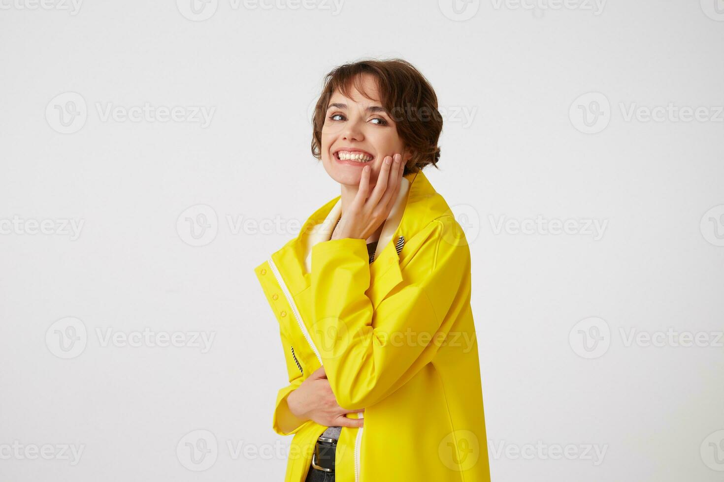 Portrait of young happy cute short haired girl wears in yellow rain coat, wbroadly smiles and looks at the camera, touches cheek, stands over white wall. photo