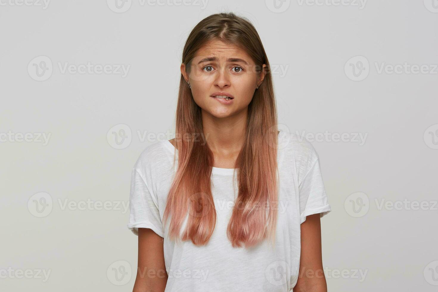 retrato de nervioso joven hermosa rubia dama con natural maquillaje mordiendo preocupantemente debajo del labio mientras mirando emocionalmente a cámara, en pie en contra blanco antecedentes foto