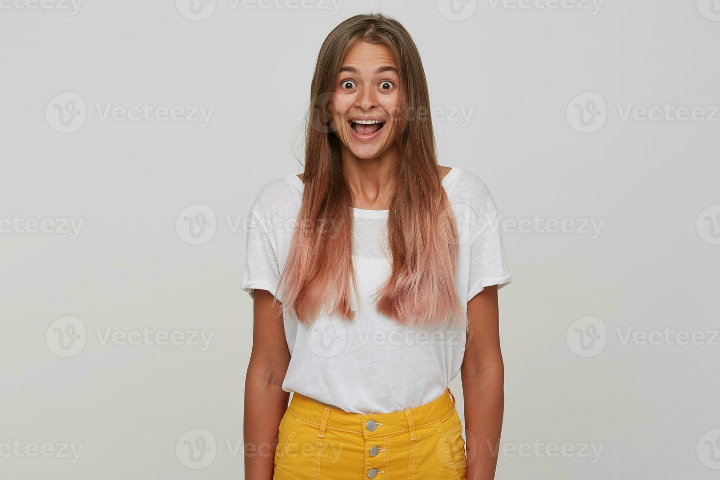 Amazed young attractive long haired blonde lady looking at camera with wide eyes and mouth opened, keeping hands along her body while standing over white background photo